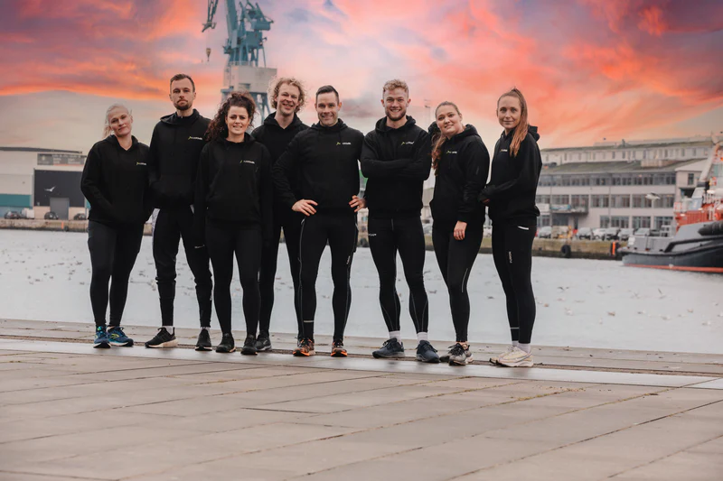 Ni personer i sort sportstøj står ved en havn ved solnedgang.